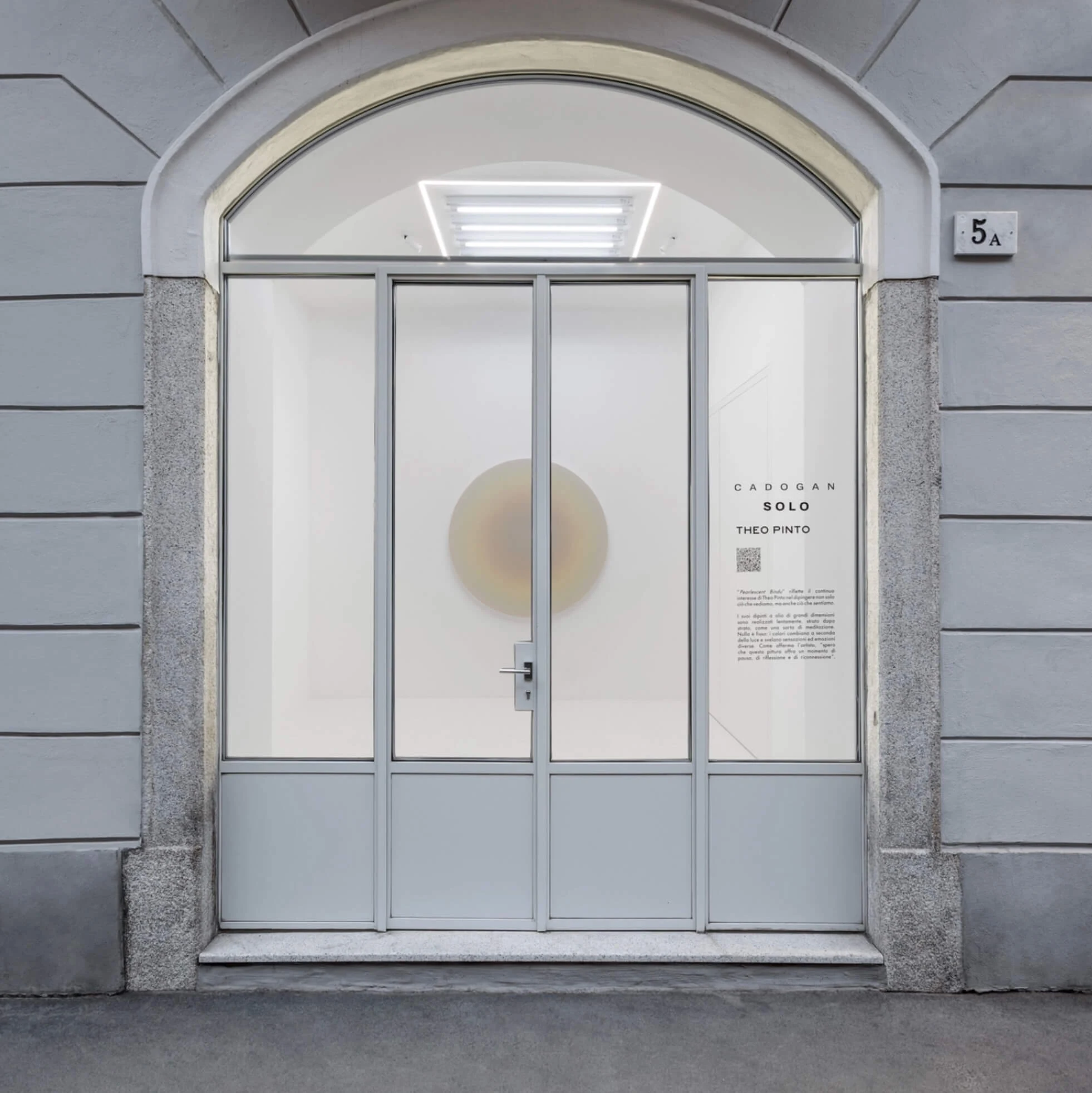 A gallery entrance seen from the street, arched glazing above the doors and a pale circular painting hung inside.